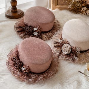 Puede incluir: Tres sombreros pequeños, redondos y de copa plana en tonos rosa empolvado y crema. Cada sombrero presenta un ala ancha de encaje con volantes, una cinta marrón oscura y un adorno de rosa de tela con detalles de encaje blanco en el lateral.