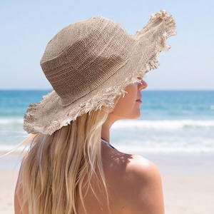 Puede incluir: Un sombrero de sol de ala ancha hecho de un material tejido texturizado de color marrón claro. El sombrero presenta una copa redondeada y un ala flexible e irregular con un acabado de borde deshilachado que crea un aspecto casual y desgastado.