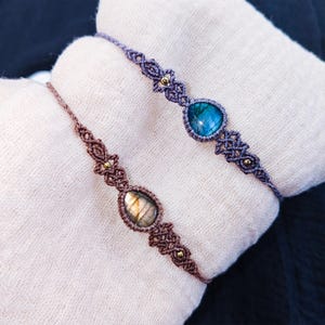Puede incluir: Dos pulseras de macramé con piedras preciosas en el centro. Una pulsera es marrón con una piedra preciosa marrón claro, y la otra es morada con una piedra preciosa azul. Ambas pulseras tienen detalles dorados.