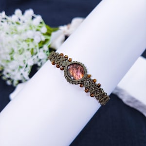 May include: A handcrafted macrame bracelet featuring a central, round, iridescent gemstone in shades of purple and pink. The bracelet is olive green with small amber-colored beads. The bracelet is displayed on a white surface.