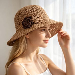 May include: A tan crocheted straw bucket hat featuring a wide brim and two decorative fabric flowers on the side, one in a dark brown shade and one in a lighter tan shade, crafted with an open-weave texture throughout the entire hat structure.