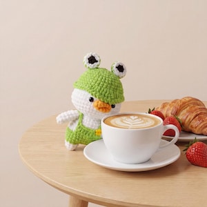 Puede incluir: Un pato de ganchillo con un gorro y un peto verdes en forma de rana. Una taza blanca de café con leche con arte, un cruasán y fresas están sobre una mesa de madera. El pato tiene un pico amarillo y ojos negros.