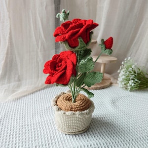 Può includere: Una pianta di rose all'uncinetto fatta a mano con due rose rosse vibranti, foglie verdi e un bocciolo, il tutto in un vaso beige. Il vaso ha un bordo decorativo e una base marrone. La pianta è su una superficie bianca.