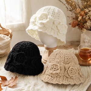 May include: Three crochet bucket hats in white, black, and beige. Each hat features a textured knit pattern and a band of three-dimensional crocheted flowers around the crown. The hats have a rounded top and a short, slightly flared brim.