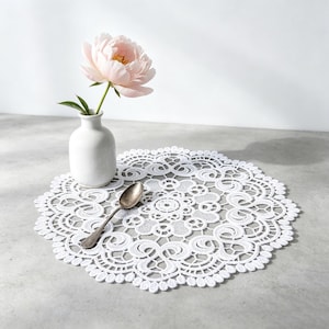 Puede incluir: Un mantel individual redondo de encaje blanco con un intrincado diseño floral. Un pequeño jarrón blanco contiene una peonía rosa. Una cuchara plateada descansa sobre el mantel individual. Los elementos están dispuestos sobre una superficie gris, creando una naturaleza muerta delicada y elegante.