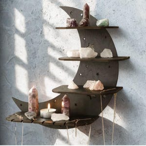 May include: A dark brown, crescent moon-shaped shelf with three tiers, displaying crystals and a lit candle. The shelf features star cutouts and hanging necklaces. Crystals are in shades of purple, pink, green, and white. The shelf is mounted on a wall.