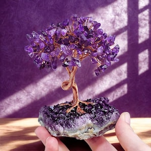 Könnte beinhalten: Eine handgefertigte Amethyst-Baumskulptur. Der Baum hat einen Stamm und Äste aus Kupferdraht, verziert mit Amethyst-Edelsteinblättern. Die Basis ist eine grob geschnittene Amethyst-Geode, die eine natürliche und künstlerische Darstellung schafft.