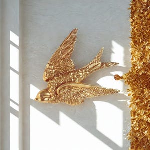 Puede incluir: Una escultura de pared dorada con forma de golondrina, con las alas extendidas. El ave está montada en una pared blanca texturizada, con sombras proyectadas por la luz solar. La escultura presenta detalles de plumas y una forma curvada.