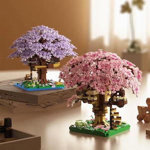 May include: Two miniature tree models, one with purple blossoms and the other with pink blossoms. The trees are constructed from small building blocks, with brown trunks and green bases. The purple tree sits on a blue base, while the pink tree has a green base.