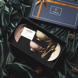 Könnte beinhalten: Ein Geschenkset mit Kaffeetassen in einer Box. Das Set enthält zwei Tassen, zwei Löffel und zwei Untertassen. Die Tassen und Untertassen haben ein Marmormuster. Die Löffel haben verzierte Griffe. Die Box ist marineblau mit einem goldenen Band und den Worten "gift & coffee cup".