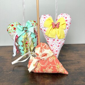 May include: Three heart-shaped fabric ornaments. One is light blue with a bow and key charm, another is white with a yellow bow and butterfly, and the third is orange with a floral pattern and bow. All have hanging ribbons.