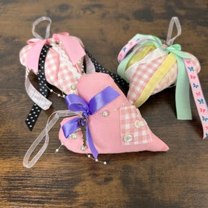 May include: Three heart-shaped ornaments with ribbons and embellishments. The hearts are pink and gingham, adorned with pearls, bows, and decorative keys. Ribbons in various colors, including pink, black, and green, add to the festive design.