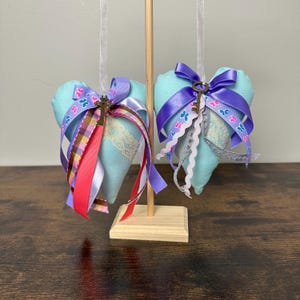May include: Two light blue heart-shaped ornaments with decorative ribbons and a small key charm. The ribbons include purple, pink, and patterned butterfly designs. The ornaments are suspended from a wooden stand, creating a charming display.