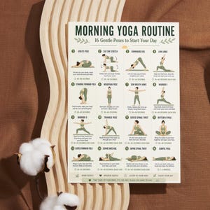 May include: A white poster titled "MORNING YOGA ROUTINE" with 16 gentle poses to start your day. The poster features illustrations of yoga poses with instructions and time durations. The poster is on a light brown surface.