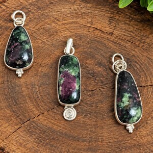 Puede incluir: Tres colgantes con piedras de rubí y zoisita pulidas engastadas en plata. Cada colgante tiene una forma única, incluyendo dos formas de lágrima y una rectangular, todas rematadas con una anilla de plata y adornadas con pequeños detalles.