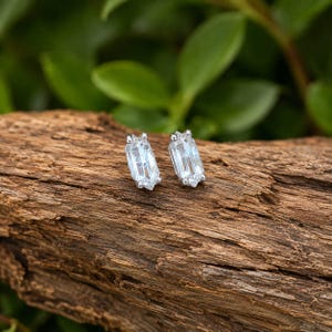 Peut inclure: Une paire de boucles d'oreilles à clous avec des pierres transparentes taille baguette serties dans un métal argenté. Chaque boucle présente une forme rectangulaire allongée aux surfaces facettées qui reflètent la lumière, fixée par un sertissage à griffes sur une surface en bois texturé.