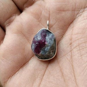 Puede incluir: Colgante de rubí natural con colgador de plata. La piedra preciosa es una mezcla de tonos rojo intenso y grises, engastada en un marco de plata pulida y sencilla. Una pieza de joyería única.