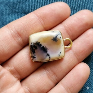 May include: A square pendant with a gold-colored frame and a loop. The pendant features a cream-colored stone with black and brown dendritic inclusions, resembling a landscape. The pendant is held in a hand.