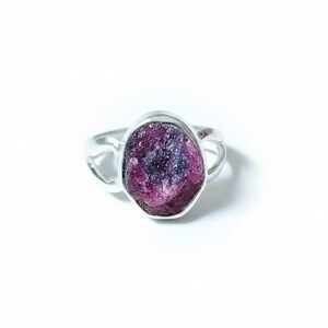 Puede incluir: Anillo de plata con una gema grande, texturizada y de color púrpura intenso. La gema presenta tonos de rosa y violeta. El diseño del anillo es sencillo y abierto.