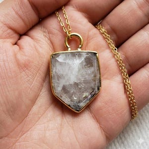 May include: A gold-colored necklace featuring a shield-shaped pendant with a clear, textured stone. The stone has white and gray internal patterns. The necklace chain is also gold-colored.