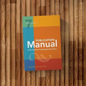以下が含まれることがあります： 「Publication Manual of the American Psychological Association, Seventh Edition」というタイトルのカラフルな本。表紙は青、オレンジ、赤の縦型カラーブロックデザインです。本は木製の表面にあります。