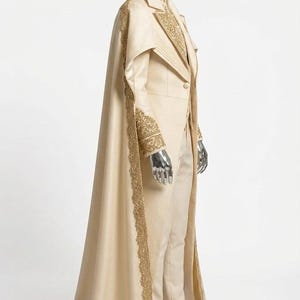 Puede incluir: Un traje color crema con una capa a juego, adornado con bordados dorados. El traje presenta una chaqueta entallada, pantalones y una larga capa que se arrastra por el suelo. La capa y la chaqueta tienen ribetes dorados.