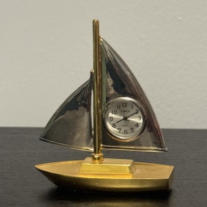 May include: A gold-toned sailboat figurine featuring a polished silver-toned sail that serves as a clock face. The clock has a white dial with black numerals and the text Timex Quartz. The boat has a gold-toned hull and a central mast.