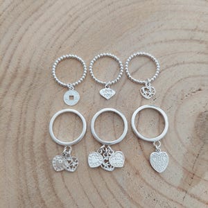 May include: Six silver rings with various charms, including a coin, a heart with the words "LOVE YOU", and heart-shaped designs. The rings are displayed on a wooden surface.