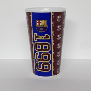 May include: A white ceramic tumbler with a maroon background and the text "1899" in gold. The tumbler features the FC Barcelona crest and repeating team logos. The design is in blue, gold, and maroon.