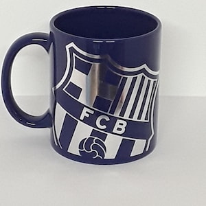May include: A navy blue ceramic mug with a white and silver emblem featuring the letters "FCB" and a soccer ball. The mug has a curved handle and a cylindrical shape.