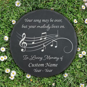 May include: A round, dark gray memorial stone with white text and musical notes. The inscription reads "Your song may be over, but your melody lives on." Below, it says "In Loving Memory of Custom Name Year - Year."
