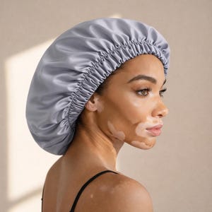 Puede incluir: Un gorro de satén gris claro con una banda elástica. El gorro está diseñado para proteger el cabello mientras se duerme. La imagen muestra el gorro en una persona con una camiseta negra.