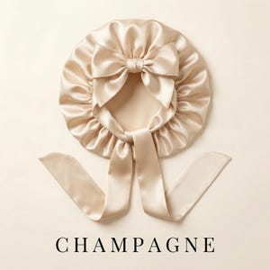 May include: A champagne-coloured satin bonnet with a large bow and long ties. The bonnet is round and has a gathered design. The word "CHAMPAGNE" is printed in black block letters at the bottom.