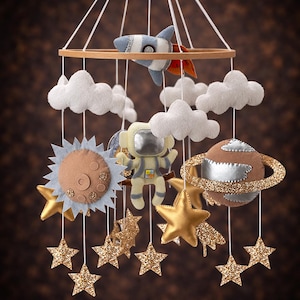 Op de afbeelding: Handgemaakte baby mobile met een houten ring en vilten ruimte-elementen. Inclusief een raket, astronaut, planeet, zon, wolken en gouden glittersterren. De mobile hangt aan witte touwtjes, perfect voor de kinderkamer.