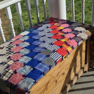 Puede incluir: Un cofre de madera con una colcha de retazos en la parte superior. La colcha presenta cuadrados de varios colores y patrones, incluyendo rojo, azul y cuadros escoceses, asegurados con lazos de hilo azul. El cofre es de madera clara.