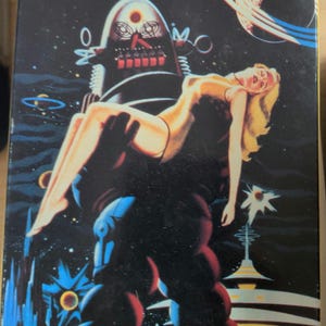 Könnte beinhalten: Eine Vintage-VHS-Kassette für "Forbidden Planet: New Remastered Edition". Das Cover zeigt eine Retro-Sci-Fi-Illustration eines Roboters, der eine Frau hält, vor einem Weltraumhintergrund. Das MGM/UA Home Video-Logo ist sichtbar.