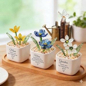 May include: Three small white ceramic pots with glass flowers in yellow, blue, and white. Each pot has a message for Mom and Grandma. The pots sit on a wooden tray with a watering can and books in the background.