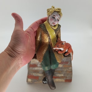 Może przedstawiać: Ceramiczna figurka przedstawiająca osobę o blond włosach w złotym płaszczu, żółtym szaliku i ciemnozielonych spodniach, siedzącą na ceglanym murku z małym pomarańczowym lisem odpoczywającym na kolanach. Figurka jest pomalowana w detale.
