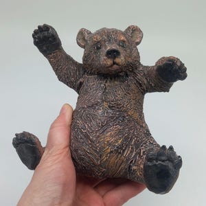 Pode incluir: Uma estatueta de urso castanho com os braços estendidos e em pose sentada. O urso tem uma superfície texturizada, patas escuras e um rosto detalhado. A estatueta é segurada em uma mão contra um fundo branco.