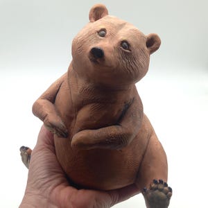 Può includere: Una statuetta di orso in argilla marrone con superficie ruvida, orecchie arrotondate e muso liscio. L'orso è raffigurato in posizione seduta con le zampe anteriori ripiegate vicino al petto e le zampe posteriori visibili alla base.