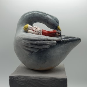 Può includere: Una scultura in ceramica di un grande cigno grigio con il becco giallo, con una piccola figura dai capelli biondi, un top bianco a motivi e pantaloni rossi che riposa sul dorso del cigno. Il cigno è posizionato su una base grigia testurizzata.
