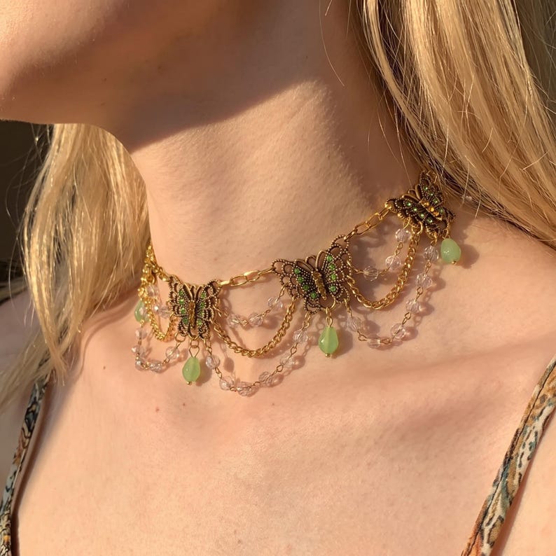 May include: A gold-toned choker necklace featuring three butterfly charms with green accents. The design includes draped gold chains and clear crystal beads, with small, light green teardrop-shaped pendants hanging from the butterfly motifs.