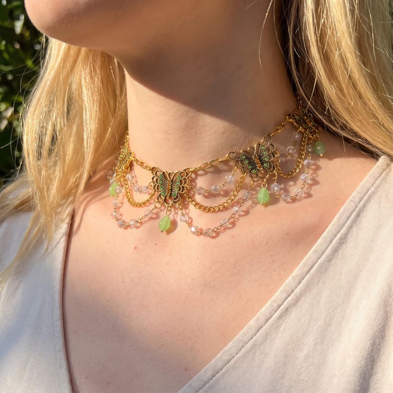 May include: A gold-toned chain choker necklace featuring multiple filigree butterfly charms with green accents. The design includes draped clear crystal beads and dangling light green teardrop-shaped beads, creating a layered, ornate aesthetic.