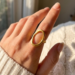 Peut inclure: Une bague dorée présentant une grande pièce centrale ovale, ouverte et irrégulière, avec une finition mate et lisse. L'anneau est simple et fin, se connectant à la base de l'imposante monture ovale creuse aux formes organiques sur le doigt.