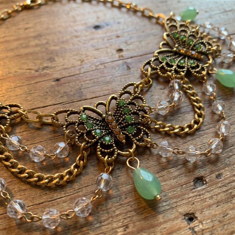May include: A gold-tone metal necklace featuring two filigree butterfly charms embellished with small green and amber-colored crystals. The design includes draped gold chains accented with clear faceted beads and a single green teardrop pendant.