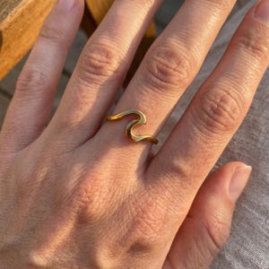 Può includere: Un anello color oro caratterizzato da un design continuo e liscio a forma di onda che avvolge il dito. Il metallo presenta una superficie lucida e riflettente che cattura la luce, creando una silhouette semplice e fluida sulla mano.