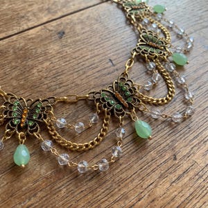 May include: A gold-toned metal necklace featuring multiple butterfly-shaped charms with green and orange accents. The design includes draped gold chains, clear faceted beads, and dangling light green teardrop-shaped stones throughout.