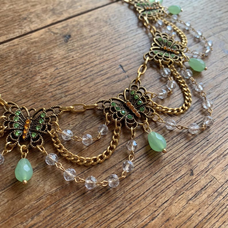 May include: A gold-toned metal necklace featuring multiple butterfly-shaped charms with green and orange accents. The design includes draped gold chains, clear faceted beads, and dangling light green teardrop-shaped stones throughout.