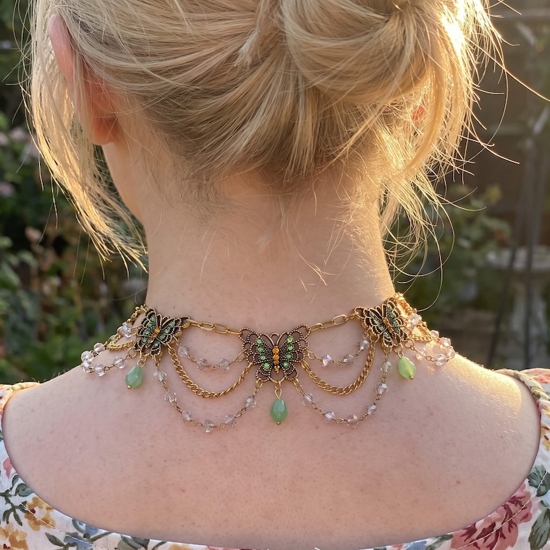 May include: A gold-toned necklace featuring three butterfly charms with green and orange accents. The piece includes draped gold chains and clear crystal beads, with three dangling light green teardrop-shaped stones suspended from the butterfly charms.