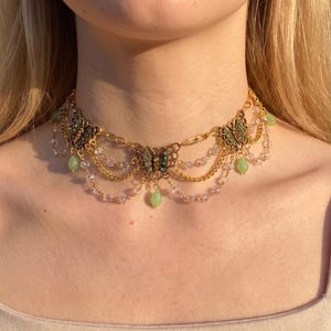 May include: A gold-toned choker necklace featuring three butterfly charms with green accents. The design includes delicate gold chains draped between the butterflies, interspersed with clear beads and small, dangling light green teardrop-shaped stones.